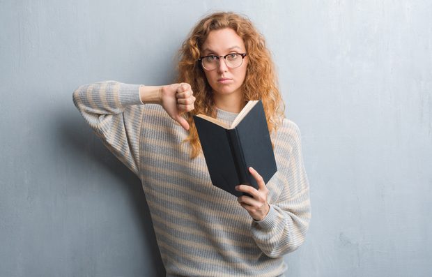 Young redhead woman over grey grunge wall reading a book with angry face, negative sign showing dislike with thumbs down, rejection concept