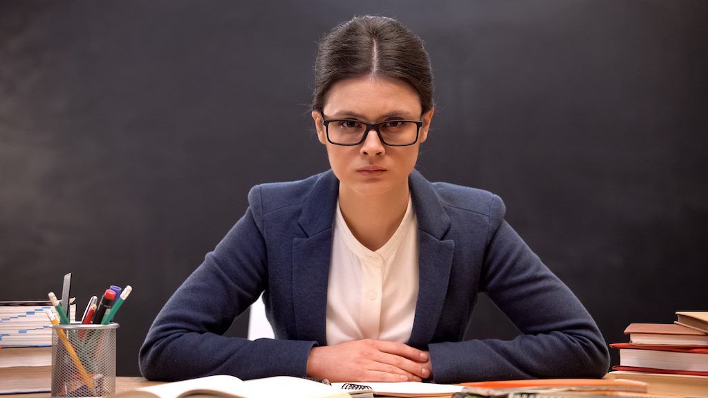 Serious head teacher aggressively looking to camera sitting by table, education