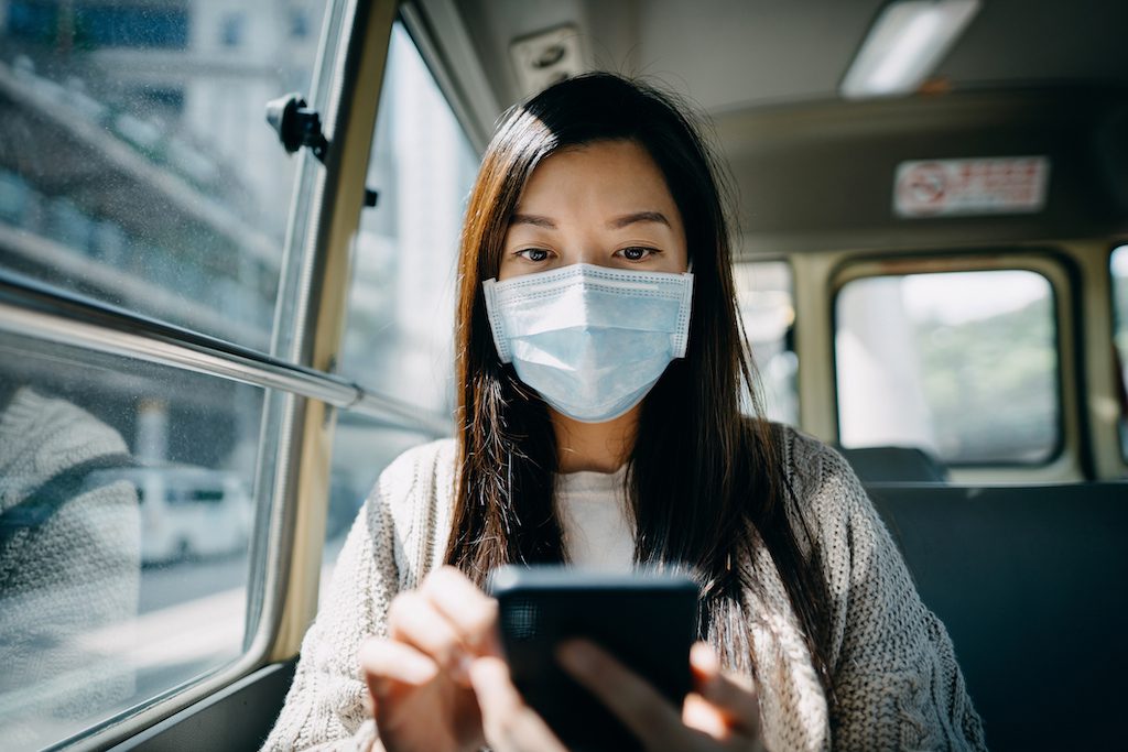 Young Asian woman with protective face mask using smartphone while commuting in the city riding on a minibus