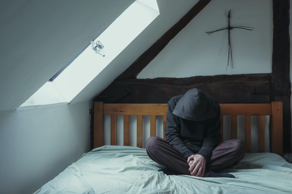 A hooded figure sitting on a bed, looking down depressed. With a cross made of twigs above the bed
