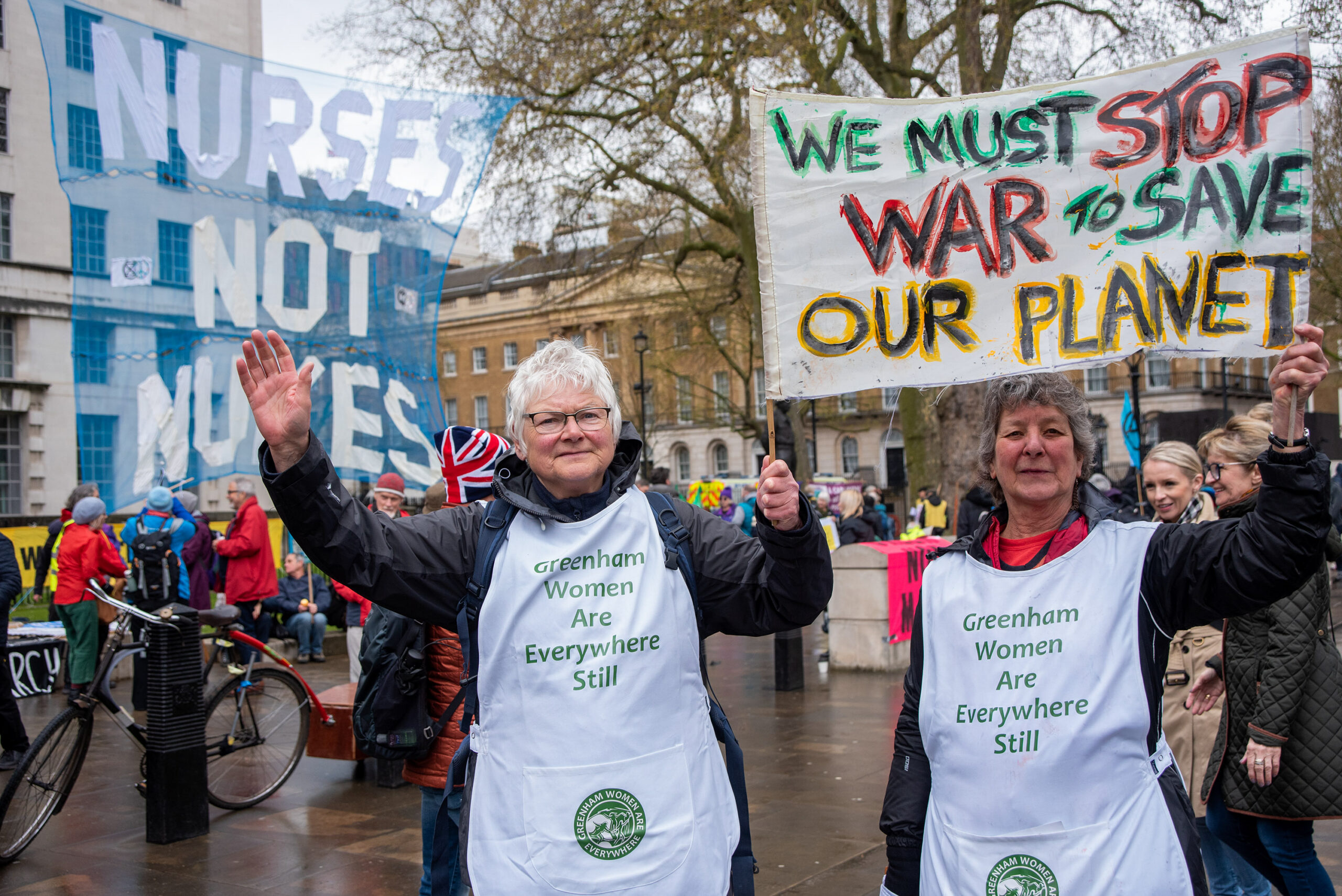Two women wearing aprons/uniforms hold a banner that says, '