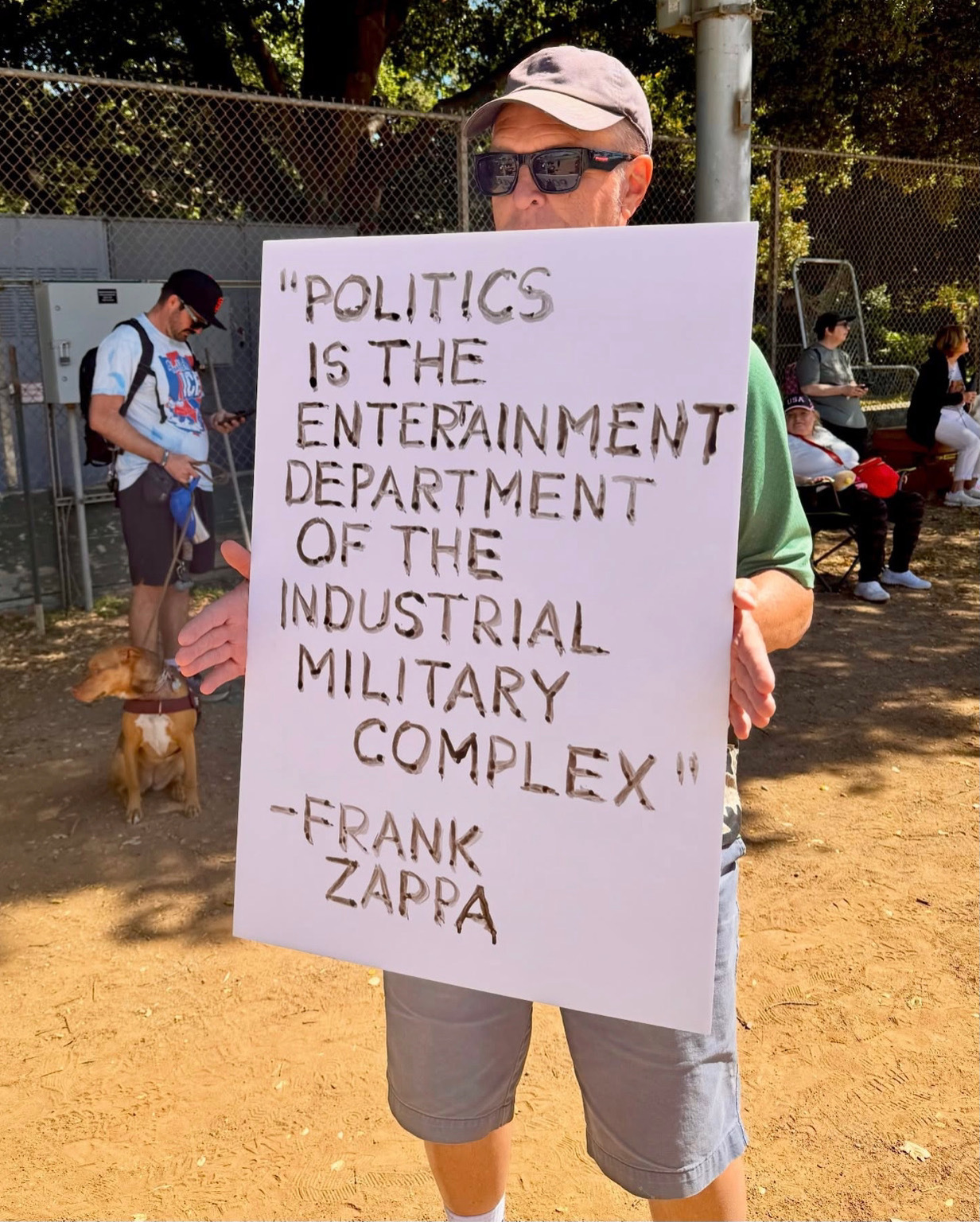 A man holding a sign with a quote from Frank Zappa: "Politics is the entertainment department of the industrial millitary complex."